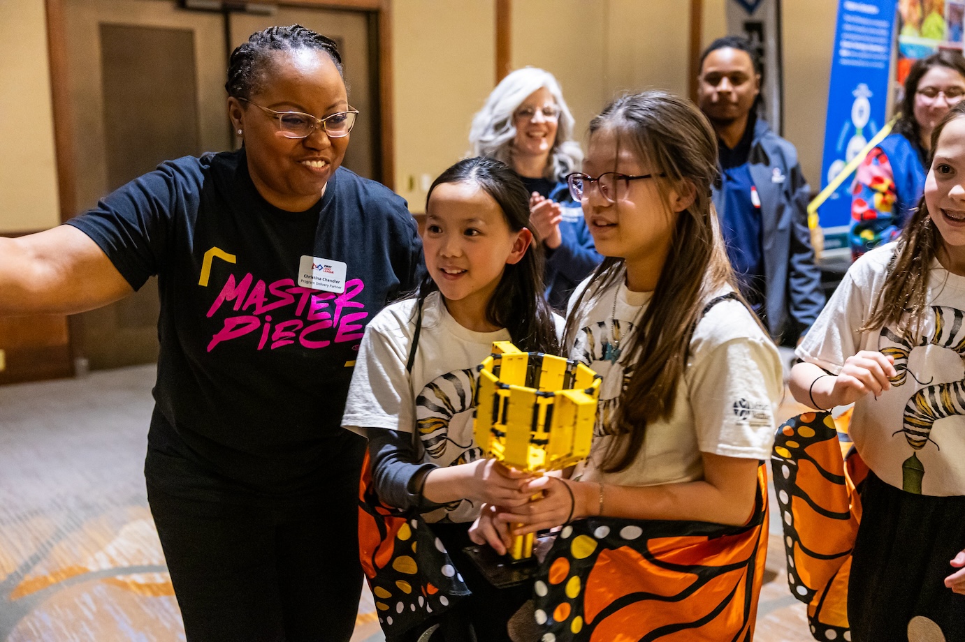 FIRST LEGO League partner Chrissy Chandler guides two girls carrying a LEGO trophy across the stage at the KC Regional Championships